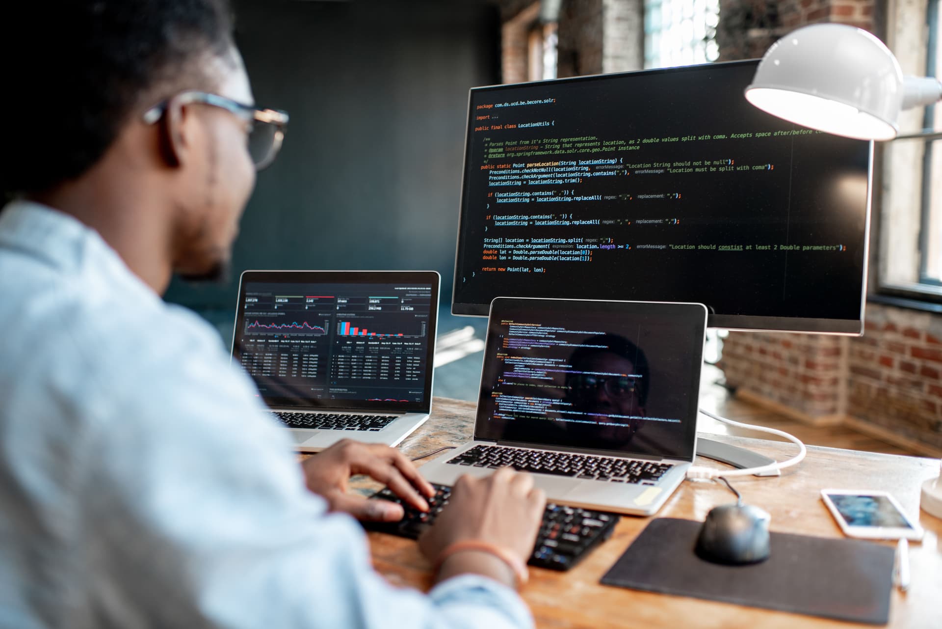 Web developer coding on three screens — two laptops and a monitor — showing code and data analytics in a modern, naturally lit office.
