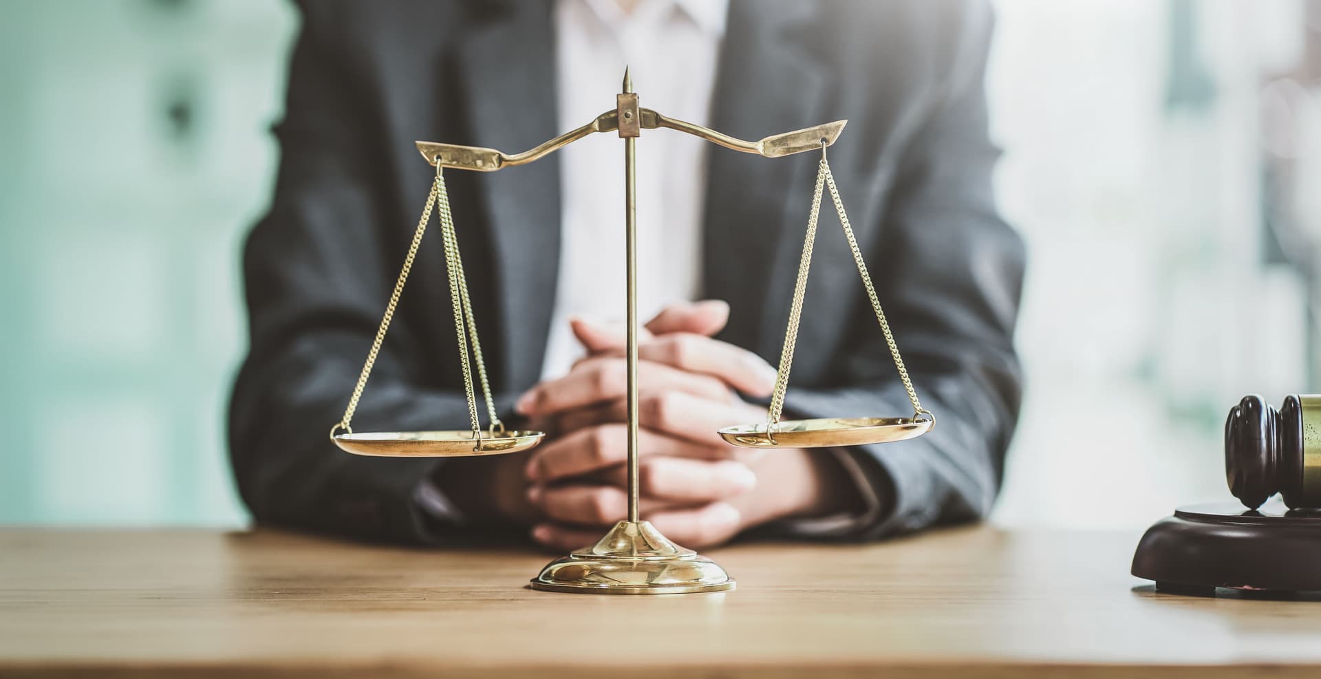 Lawyer in suit sitting behind a golden justice scale and gavel on desk, symbolizing fairness and legal decision-making.